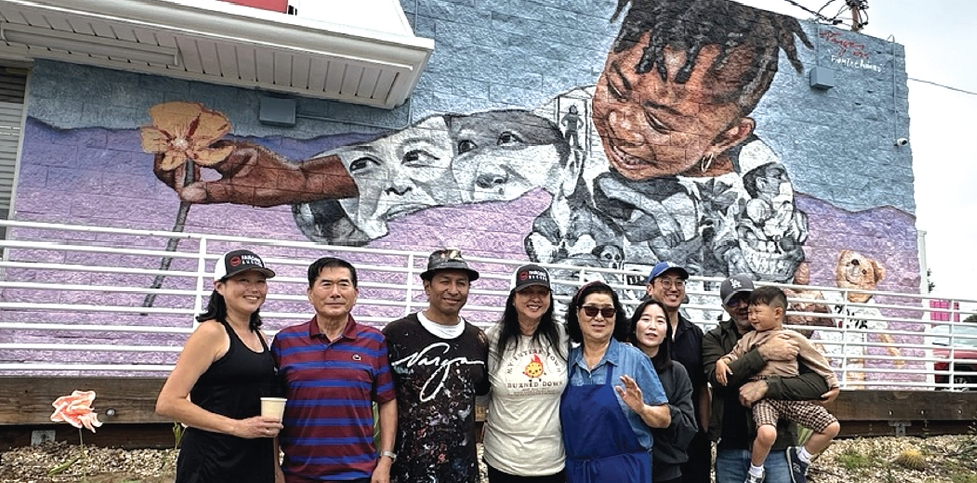 지난 7일 ‘페어옥스 버거(Fair Oaks Burger)’ 외벽에 로버트 바가스의 벽화 ‘잿더미로부터’가 공개됐다. 바가스(왼쪽 세 번째)와 창업주 이기선·유정자 씨 부부와 가족들이 벽화를 배경으로 기념사진을 찍고 있다. 김상진 기자